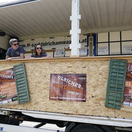 Zwei Personen sind in einem Lastwagen und schauen aus den Fenstern. Der Lastwagen hat ein hölzerne Außenwand mit einem Schild, das 'Ulricher Hutten Gaudi Warm Up' steht. Es gibt Plakate an der Seite.