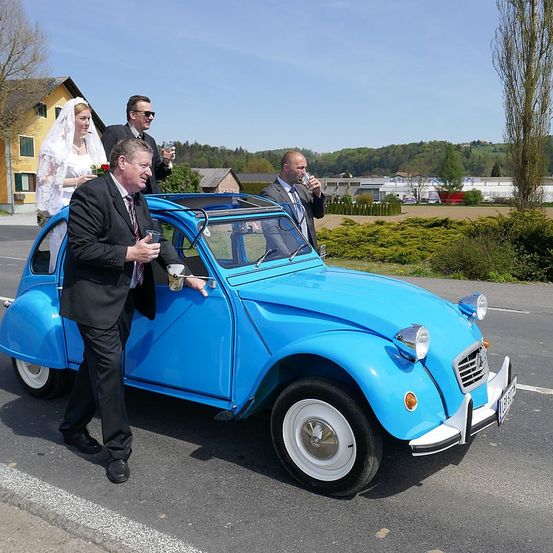 Eine Hochzeitsfeier in einem blauen Oldtimer, mit der Braut auf dem Dach, und drei Männer stehen um das Auto herum. Der Mann auf der linken Seite hält ein Getränk, während die anderen beiden Männer trinken.