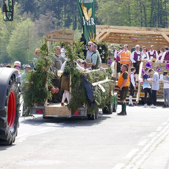 Ein Festwagen fährt auf einer Straße mit Menschen in traditioneller Kleidung. Der Wagen ist mit Grünpflanzen und einem Banner geschmückt. Zuschauer stehen am Straßenrand und beobachten. Bäume und eine Holzkonstruktion sind im Hintergrund.