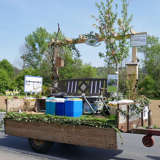 Ein Anhänger mit einer Bank und Eimern auf der Straße. Ein Schild befindet sich auf der linken Seite und ein Baum mit Blumen hinter der Bank. Eine Holzstruktur befindet sich rechts.