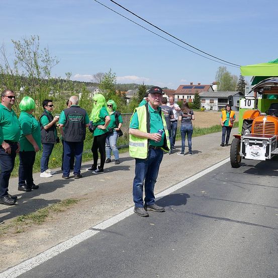 Eine Gruppe von Menschen in grünen Shirts und Warnwesten steht am Straßenrand, mit einem Traktor in der Nähe. Ein Mann spricht, während andere zuhören, in der Nähe eines Grasbereichs und Gebäude in der Ferne.