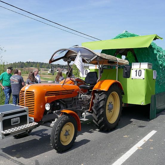Ein orangefarbener Traktor mit einem grünen Anhänger ist auf der Straße geparkt, mit einer weißen Flagge befestigt. Einige Leute stehen hinter dem Traktor.