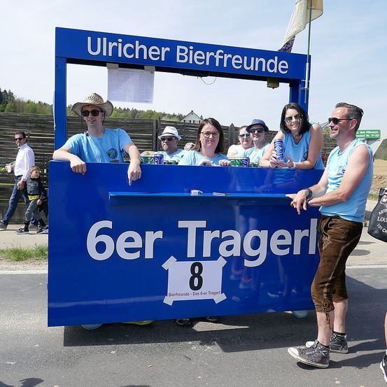 Eine Gruppe von Menschen steht hinter einem blauen Tresen mit den Worten 'Ulricher Bierfreunde 6er Tragerl' und der Nummer 8. Sie tragen blaue Hemden. Ein Mann steht rechts und trägt eine Sonnenbrille und braune Hosen.