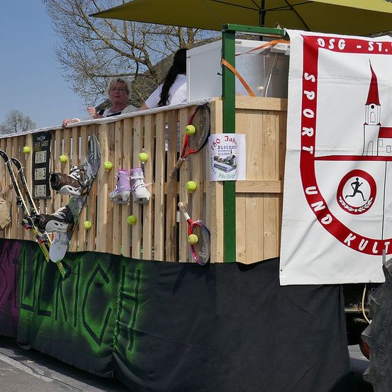 Ein Festwagen mit einem Banner, das 'Sport und Kult' besagt. Eine Frau und ein Kind stehen darauf, umgeben von Sportausrüstung.