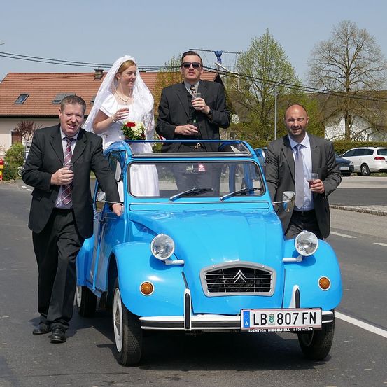 Ein frisch verheiratetes Paar, in traditioneller Hochzeitskleidung gekleidet, steht auf einem blauen Oldtimer. Das Auto ist am Straßenrand geparkt, mit Häusern und Bäumen im Hintergrund.
