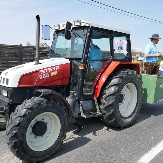 Ein roter und weißer Traktor mit der Nummer 958 ist auf der Straße geparkt. Ein Mann sitzt auf dem Fahrersitz. Ein Mann in einem Hut steht neben dem Traktor.