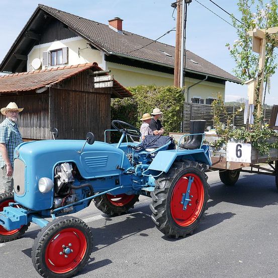 Ein blauer Vintage-Traktor mit roten Rädern steht auf der Straße vor einem Haus. In der Nähe stehen Leute, und ein hölzerner Anhänger mit der Nummer 6 ist am Traktor befestigt.