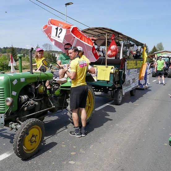 Ein Traktor zieht einen Wagen mit Leuten darauf. Ein Mann und ein Kind sitzen im Traktor, während ein Mann daneben steht. Ein roter Regenschirm ist über ihnen.