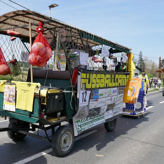 Ein grüner Wagen mit Dach und Netz, der ein 'fussballcamp'-Banner zeigt. Er enthält Sportgeräte, einen Generator und Werbematerialien. Leute gehen in der Nähe an einem sonnigen Tag.