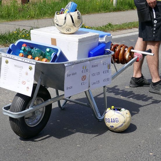 Ein Schubkarren gefüllt mit Flaschen, einem Fußball und Brezeln. Ein weißer Kühler und ein blauer Kühler sind oben darauf platziert. Eine Person in Turnschuhen steht neben dem Schubkarren.