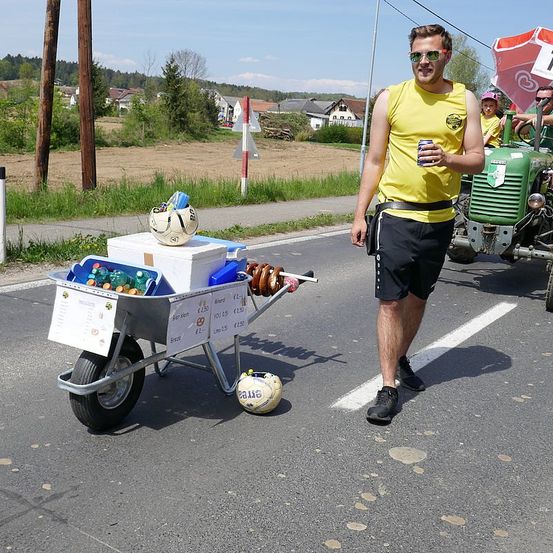 Ein Mann in einem gelben Tanktop und Sonnenbrille geht auf der Straße und schiebt einen Schubkarren voller verschiedener Gegenstände. Hinter ihm fährt ein grüner Traktor vorbei.