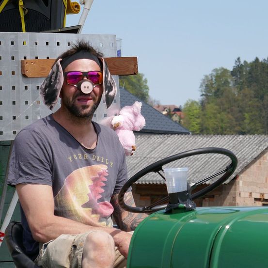 Ein Mann sitzt auf einem grünen Traktor, trägt eine Schweinemaske und ein rosa Plüschtier. Er hat Tätowierungen am Arm und trägt ein graues T-Shirt mit einem grafischen Design.