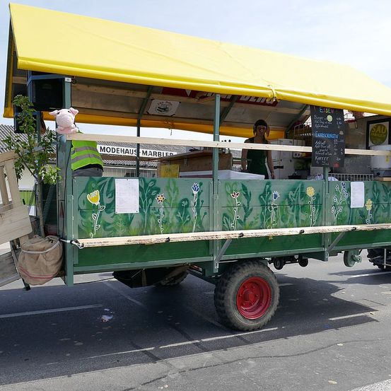 Ein grüner Imbisswagen mit gelbem Dach steht auf der Straße. Eine Seite hat einen Plüschtier, und eine Person ist drinnen. Eine Tafel mit Text steht an der Seite des Wagens.