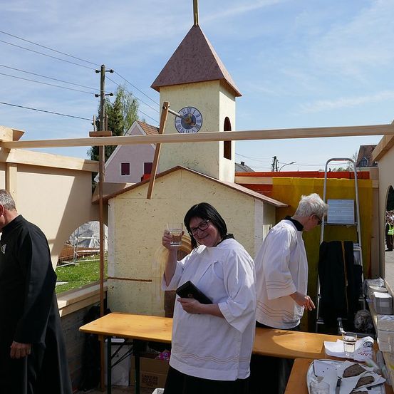 Drei Personen in weißen Gewändern stehen in der Nähe einer provisorischen Kapelle mit einem Glockenturm. Eine Frau lächelt, hält ein Glas und ein Buch. In der Nähe steht ein Mann in einem schwarzen Gewand, und ein Tisch hat Teller mit Essen.