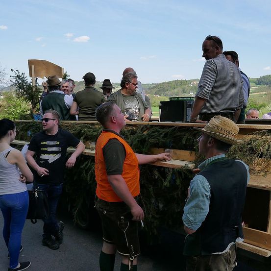 Mehrere Menschen versammeln sich um einen Holztresen, möglicherweise für ein Festival. Einige sind in traditioneller deutscher Kleidung, und ein Mann hält ein Glas. Der Hintergrund umfasst Grünpflanzen und Berge.