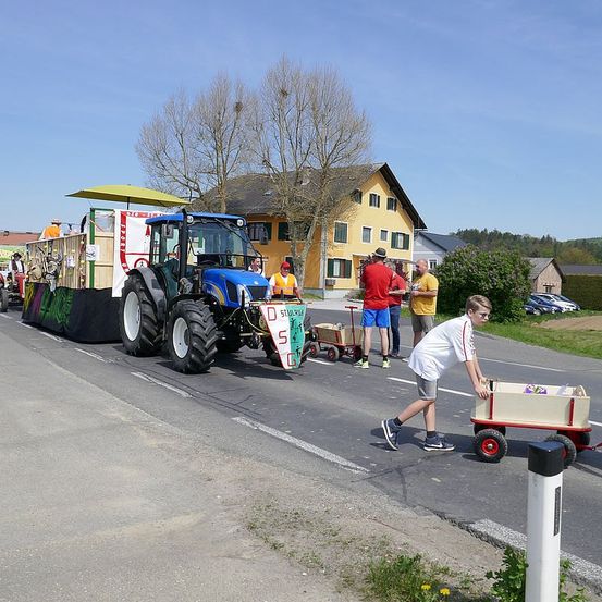 Ein Junge schiebt einen Wagen auf einer Straße während einer Parade mit einem dekorierten Traktor und Menschen im Hintergrund.