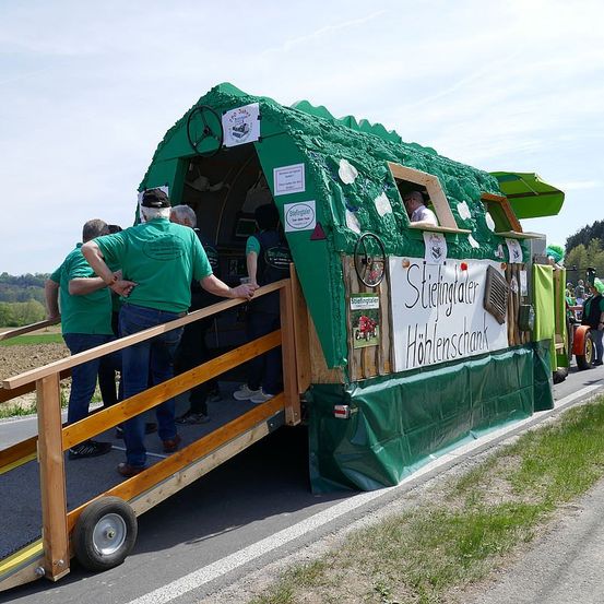 Ein grüner Festwagen mit einer Holzrampe, Leute in grünen T-Shirts und einem Banner mit der Aufschrift 'Stiftinger Hof' steht am Straßenrand.