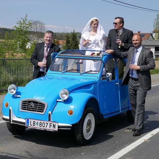 Eine Braut steht mit einem Blumenstrauß auf einem blauen Auto, während ein Bräutigam und zwei Männer aus Gläsern trinken. Sie sind draußen an einem sonnigen Tag.