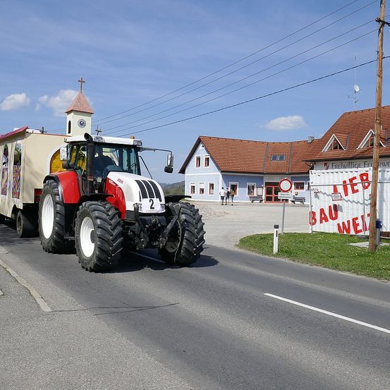 Ein dekorierter Traktor mit der Nummer 2 fährt an einem Gebäude mit dem Schild 'LIEB BAU WEI' vorbei.
