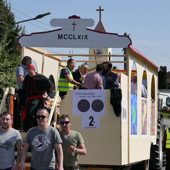 Ein Umzugswagen in einer Parade zeigt eine kirchenähnliche Struktur mit einem Kreuz oben und einem Schild, das MCCLXIX liest. Menschen sind auf dem Wagen, einige mit Sonnenbrille. Ein Mann mit rotem Hut und ein anderer in gelber Weste sind ebenfalls anwesend.