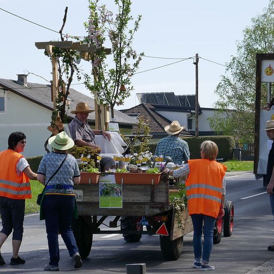 Ein Mann sitzt auf einem geschmückten Wagen mit Blumen und Pflanzen, während mehrere Menschen in orangenen Westen ihm helfen. Der Wagen steht auf einer Straße mit Häusern und Grünpflanzen im Hintergrund.
