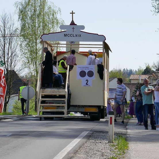 Ein Umzug mit einer kirchenähnlichen Struktur auf einem Lastwagen, die MCCLXIX anzeigt, mit Personen in Roben und Gewändern auf den Stufen. Menschen gehen am Straßenrand, einer trägt eine Warnweste. Bäume und Gebäude im Hintergrund.
