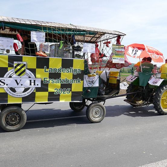 Ein Traktor mit einem Anhänger, der mit einem Netz bedeckt ist, fährt die Straße entlang, begleitet von einem Banner und einer Gruppe von Menschen.