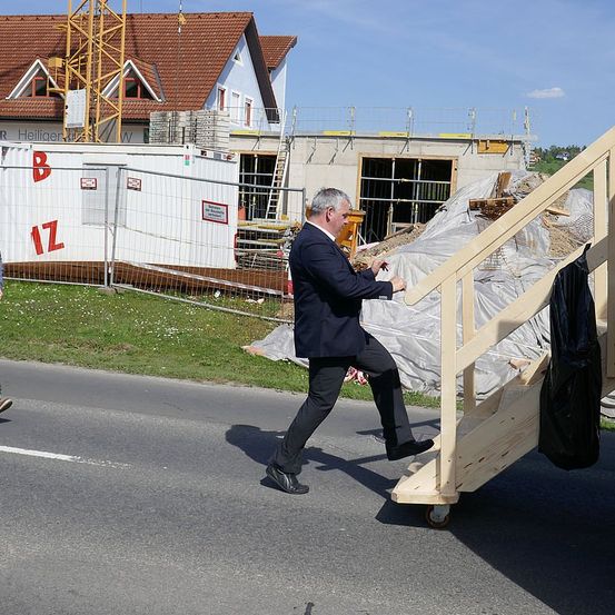 Ein Mann in einem Anzug geht eine Holzrampe zu einem Bauplatz hinauf, mit einem Haus und Maschinen im Hintergrund.