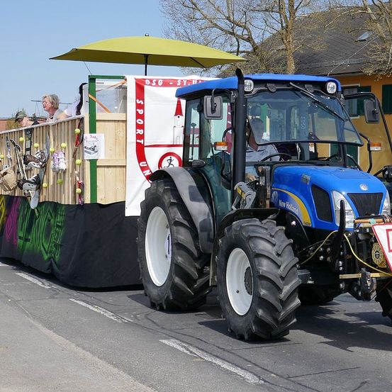 Ein blauer Traktor mit gelbem Sonnendach fährt eine Straße entlang und zieht einen Wagen mit einem Banner und winkenden Personen. Der Traktor hat große Reifen und eine weiße Felge. Der Wagen hat Tennisbälle und Skier.