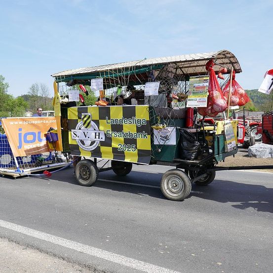 Ein gelber und schwarzer dekorierter Wagen, möglicherweise ein Festwagen, bewegt sich auf einer Straße. Er hat ein Verdeck, mehrere Bälle in roten Tüten und ein Banner, auf dem 'Landesliga Ersatzbank 2025' steht. Links befindet sich ein blaues Banner mit einem Logo und etwas Text. Ein Traktor und einige Bäume sind im Hintergrund zu sehen.