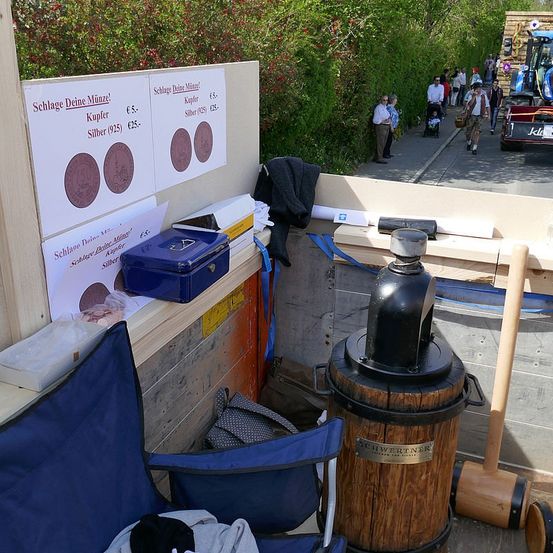 Eine Freiluftumgebung mit einem Holztisch, verschiedenen Gegenständen und einem Holzfass mit der Aufschrift Schwertner. Es gibt Münzen, eine blaue Box und einen Holzhammer. Ein Schild zeigt die Preise für Kupfer- und Silbermünzen an. Leute gehen auf dem Bürgersteig.