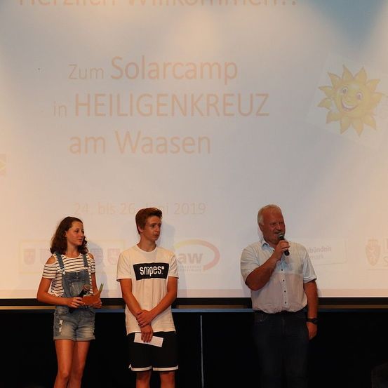 Drei Personen stehen vor einer Leinwand, die ein Solarcamp in Heiligenkreuz am Waasen vom 24. bis 26. 2019 anzeigt. Ein Mann hält ein Mikrofon und spricht. Die anderen halten Objekte.