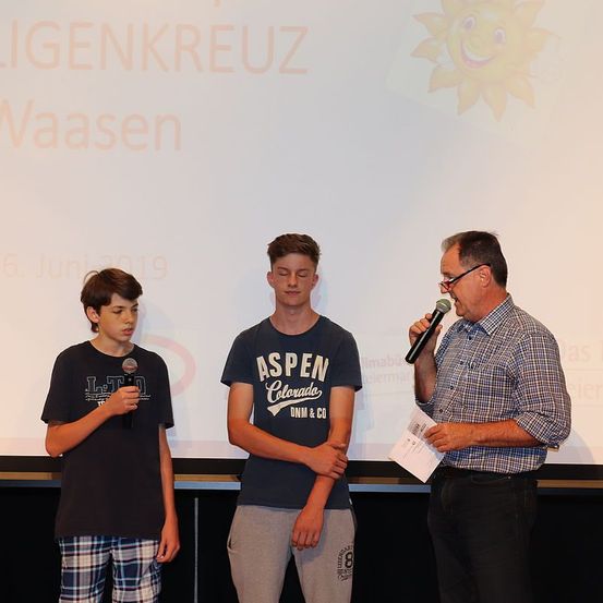 Drei Personen stehen vor einer Leinwand, wahrscheinlich bei einer Veranstaltung. Der Mann rechts hält ein Mikrofon, und die beiden Jungen neben ihm scheinen eine Auszeichnung zu erhalten.
