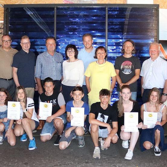 Eine Gruppe von Menschen posiert für ein Foto und hält Urkunden in der Hand. Einige tragen Brillen, Gürtel, Sneaker und Uhren. Sie stehen vor einem blauen Hintergrund.