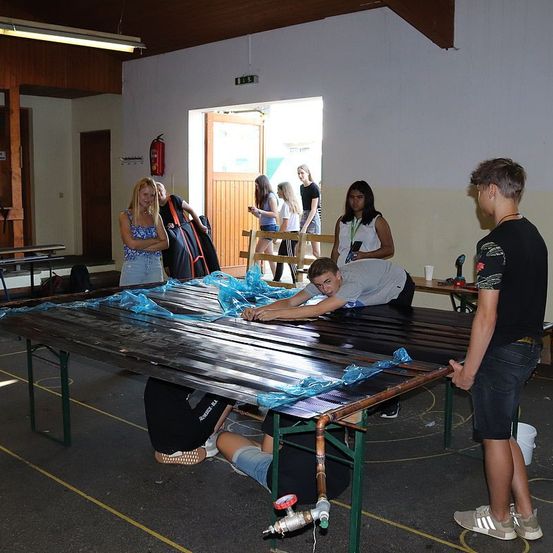Eine Gruppe von Menschen arbeitet in einem Raum an einem Tisch, an der Wand ist ein Feuerlöscher montiert. Der Tisch hat Kupferrohre und blaues Plastik. Einige Leute stehen herum.