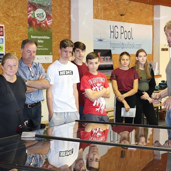 Eine Gruppe von Menschen steht um einen Glastisch herum, wahrscheinlich in einem Gebäude. Sie schauen sich den Glastisch an und unterhalten sich miteinander. Ein Banner mit den Worten 'HG Pool' ist an der Wand befestigt.