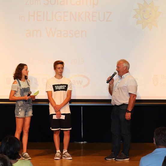 Drei Personen stehen auf der Bühne mit einem Mikrofon und einem Hintergrund. Eine Frau hält ein Papier, ein Mann ein Mikrofon und ein weiterer Mann steht zwischen ihnen.