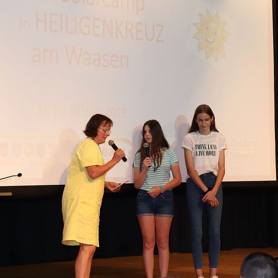 Eine Frau in einem gelben Kleid hält ein Mikrofon und spricht zu zwei Mädchen, die neben ihr auf einer Bühne vor einer großen Leinwand stehen.