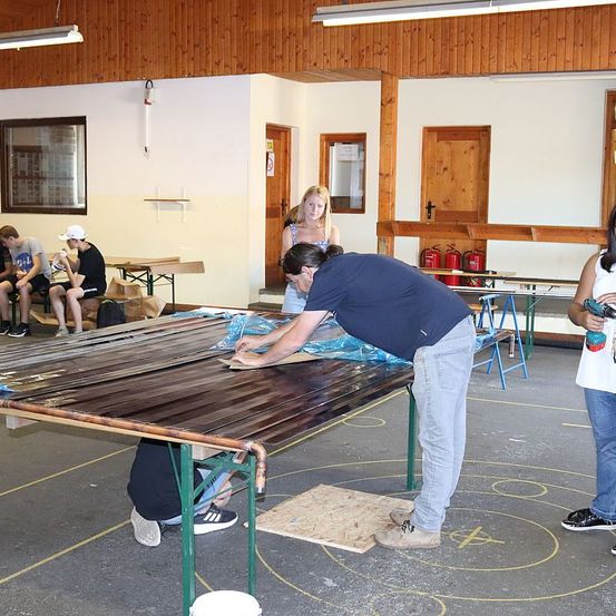 Ein Workshop mit Holzwänden, in dem ein Mann an einem Holztisch arbeitet. Zwei Personen stehen vor dem Tisch, andere sitzen auf Bänken. Ein Feuerlöscher ist an der Wand montiert.