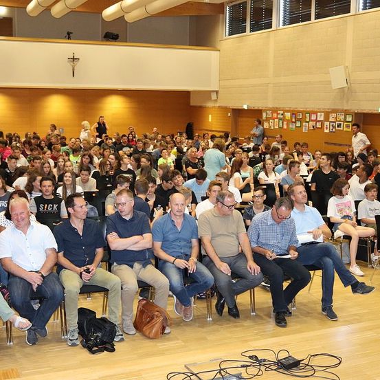Eine große Gruppe von Menschen sitzt in einem Saal, wahrscheinlich während einer Vorlesung. Einige Personen tragen Brillen, und einige haben Taschen auf den Knien. Der Saal verfügt über einen Holzboden und hohe Decken.