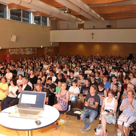 Eine große Indoor-Versammlung von Menschen, die auf Stühlen um einen Tisch mit einem Laptop sitzen. Einige klatschen in die Hände, andere schauen auf ihre Handys. Der Raum hat Holzdecken und Wände, die mit verschiedenen Bildern geschmückt sind.