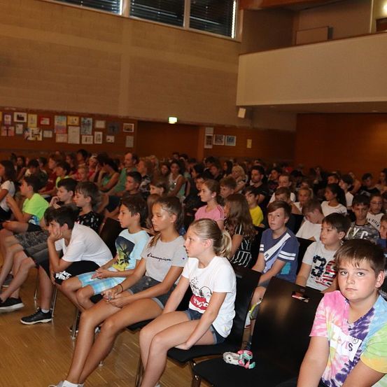 Eine Gruppe von Kindern und einigen Erwachsenen sitzt in einem Raum, möglicherweise für eine Präsentation. Einige von ihnen tragen Turnschuhe, während andere Armbänder tragen. Der Raum hat einen Holzboden und Wände, die mit verschiedenen Bildern geschmückt sind. Die Kinder scheinen auf etwas vor ihnen zu schauen.