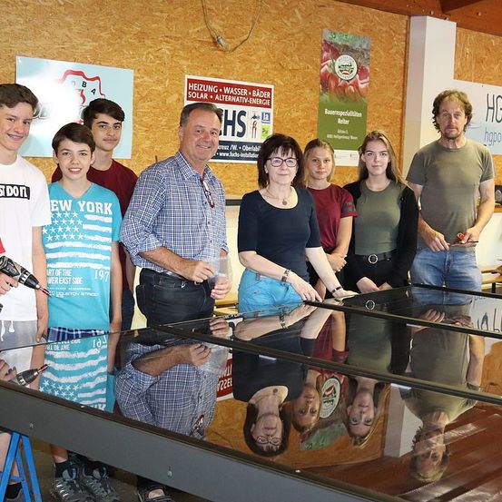 Eine Gruppe von Menschen steht hinter einem Glastisch in einem Raum mit Plakaten an der Wand. Eine Person hält eine Bohrmaschine, und die anderen schauen zur Kamera.