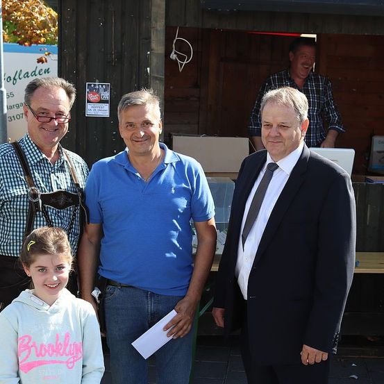 Drei Männer und ein Mädchen stehen vor einer Holzstruktur. Die Männer lächeln und scheinen für ein Foto zu posieren. Der Mann auf der rechten Seite hält ein Stück Papier. Hinter ihnen steht ein weiterer Mann in der Nähe eines Tisches.