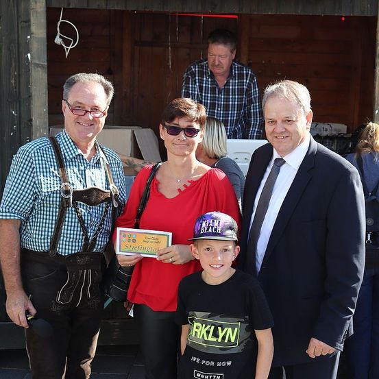Eine Familie posiert für ein Foto draußen, mit einem Mann in traditioneller bayerischer Kleidung, einer Frau, die ein Zertifikat hält, einem Jungen in einem Hut und einem Mann in formeller Kleidung. Im Hintergrund steht eine Person in der Nähe einer Holzstruktur.