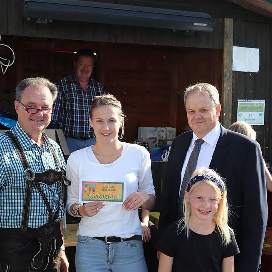 Eine Gruppe von Menschen steht in einer Scheune, eine Frau hält ein Zertifikat und lächelt. Ein älterer Mann in traditioneller deutscher Kleidung steht neben ihr. Ein anderer Mann im Anzug und Krawatte steht hinter ihnen. Ein Mädchen mit Stirnband lächelt neben ihnen. Ein Mann in einem karierten Hemd steht im Hintergrund.