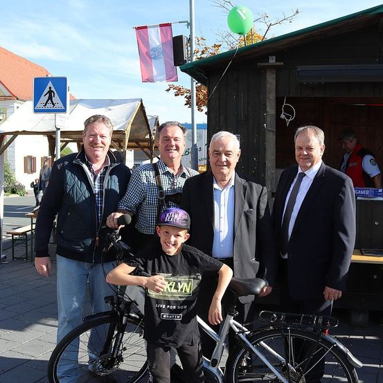 Vier Männer stehen lächelnd neben einem Jungen vor einem Fahrrad. Dahinter ist eine Holzkonstruktion mit einer Flagge und einem Schild zu sehen, und eine Person steht in der Nähe eines Tisches mit Bänken.