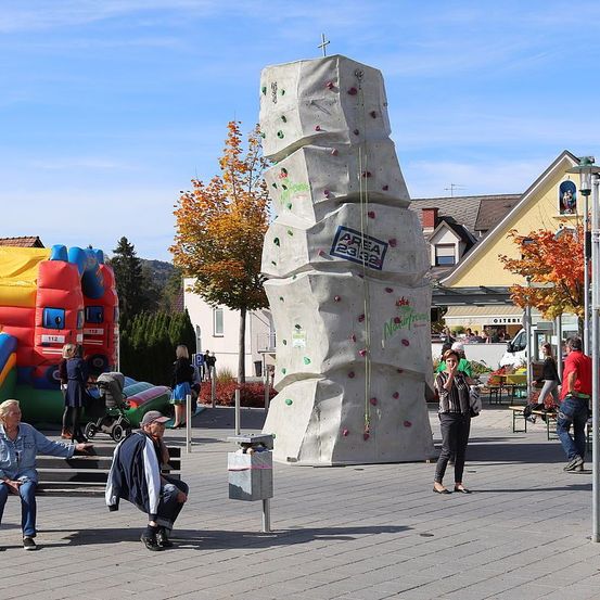 Eine große Kletterwand befindet sich in einer Freiluft-Plaza. Mehrere Menschen interagieren mit ihr. In der Nähe macht eine Frau ein Foto.