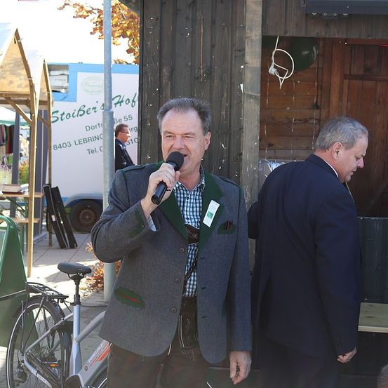 Ein Mann in einem grauen Jackett spricht in ein Mikrofon im Freien. Neben ihm steht ein weiterer Mann in einem dunklen Anzug. Dahinter befindet sich eine Holzstruktur mit einem Schild.