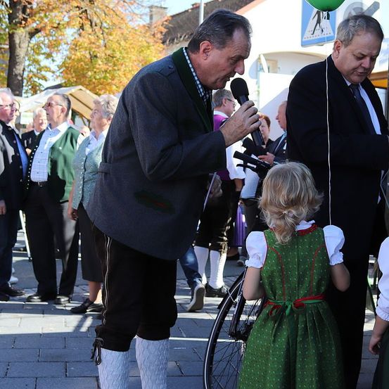 Ein Mann mit einem Mikrofon spricht zu einer Menschenmenge, wobei ein junges Mädchen in traditioneller Kleidung und ein Mann in formaler Kleidung aufmerksam zuhören. Die Szene spielt sich auf einem Außenbereich ab, wo Menschen stehen und ein Fahrrad im Vordergrund steht.